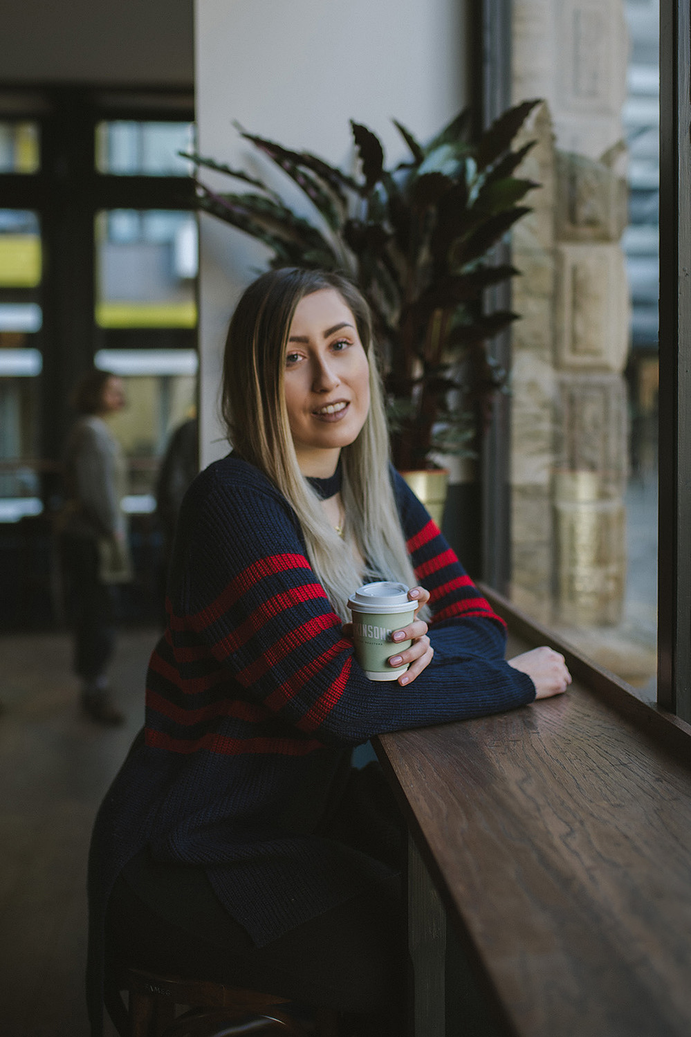 girl sits in coffee bar wearing striped jumper dress, with brown to blonde balayage hair nursing a coffee in a takeaway cup smiling at the camera