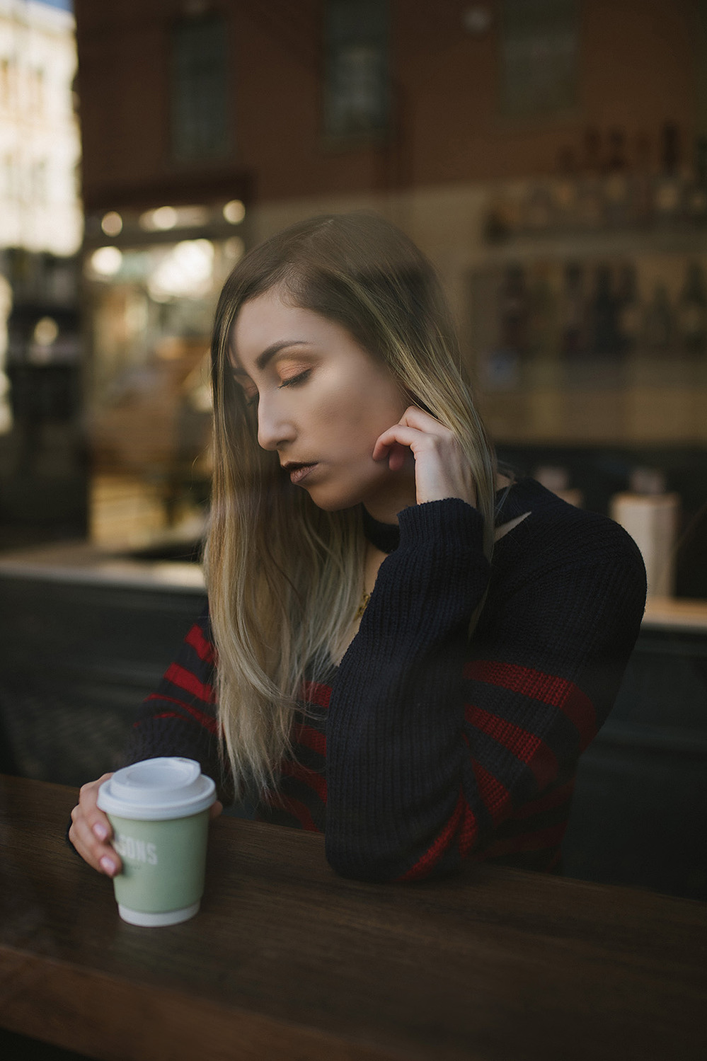 girl sits in coffee bar wearing striped jumper dress, with brown to blonde balayage hair nursing a coffee in a takeaway cup