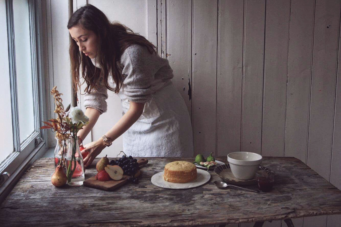 Step into the creative studio of Georgie Glass, where art meets culinary excellence. This self-portrait captures the essence of the artist at work, arranging a rustic still-life scene that whispers stories of simplicity and beauty.