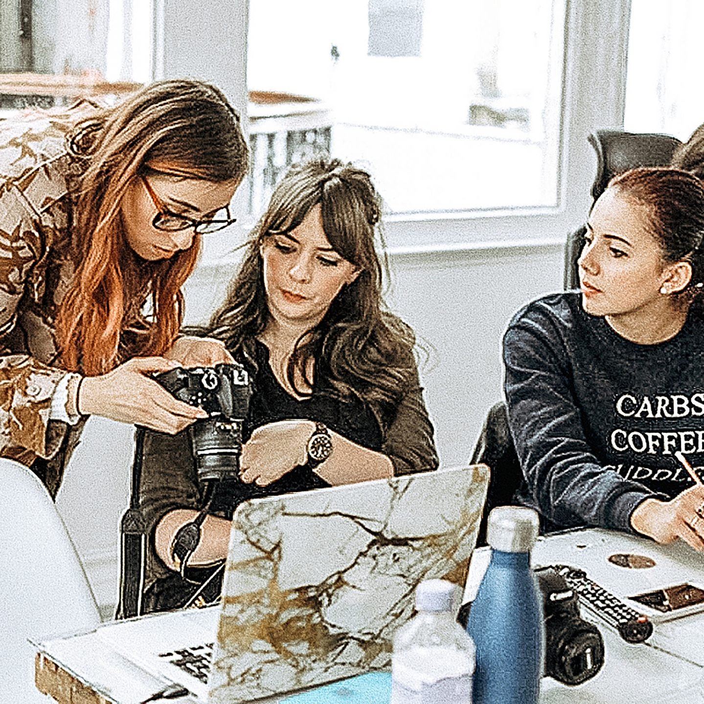 Georgie Glass photographer showing two women how to operate a camera, there's another camera, laptop and water bottle on the desk and the two women are learning about photography 