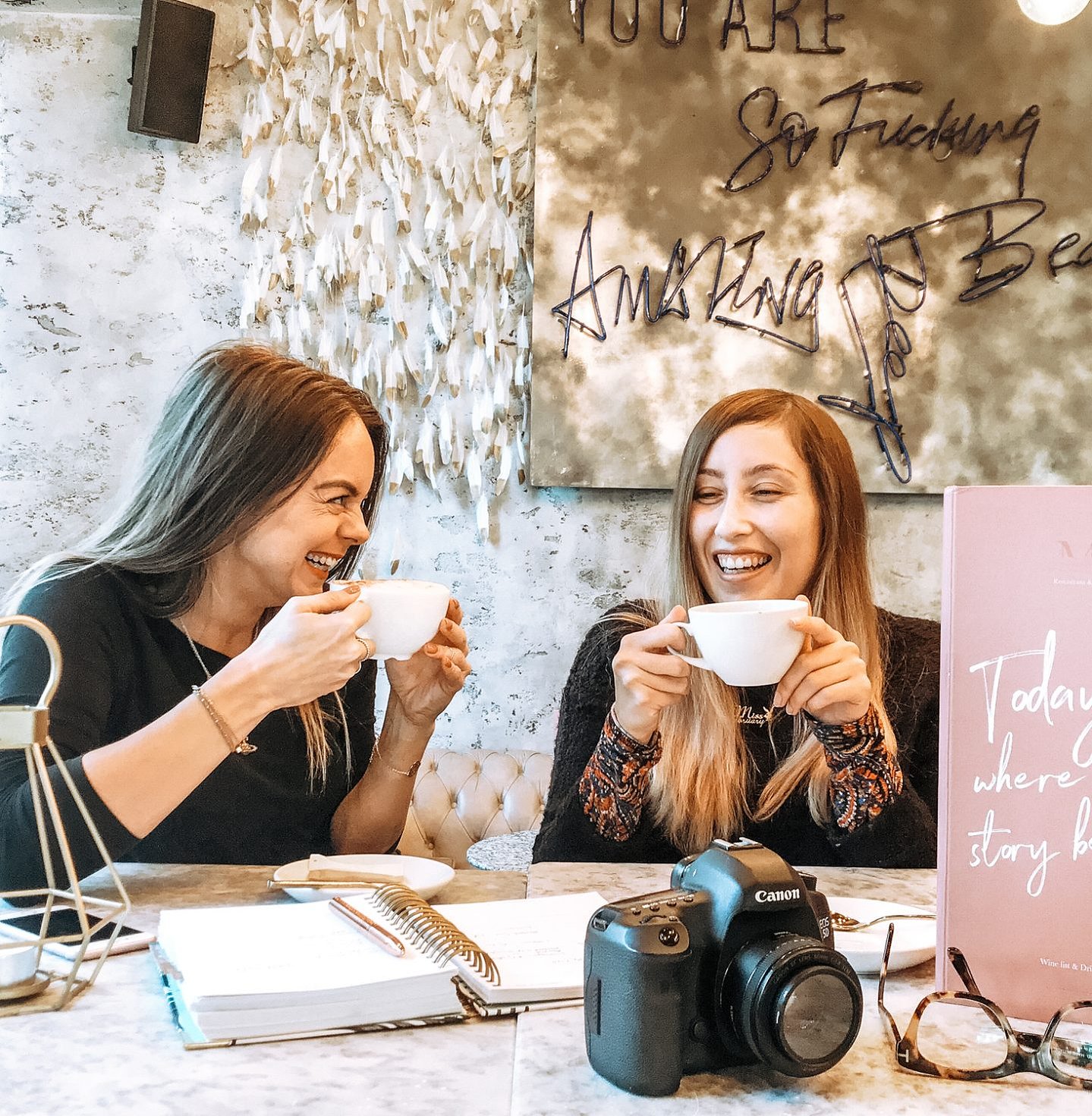 Two girls sit laughing over coffee in a restaurant with open notebooks and a Canon camera eluding that they're at a work meeting