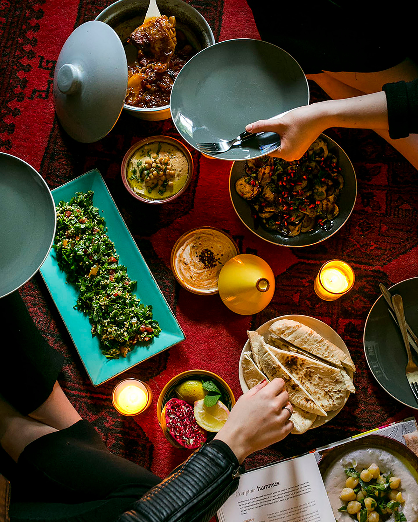 Overhead shot of a cozy Middle Eastern feast on a red patterned rug, including a tagine, hummus, tabbouleh, pita bread, and mixed vegetables, with hands serving food, photographed by Georgie Glass from Manchester, UK, capturing the warmth of shared dining
