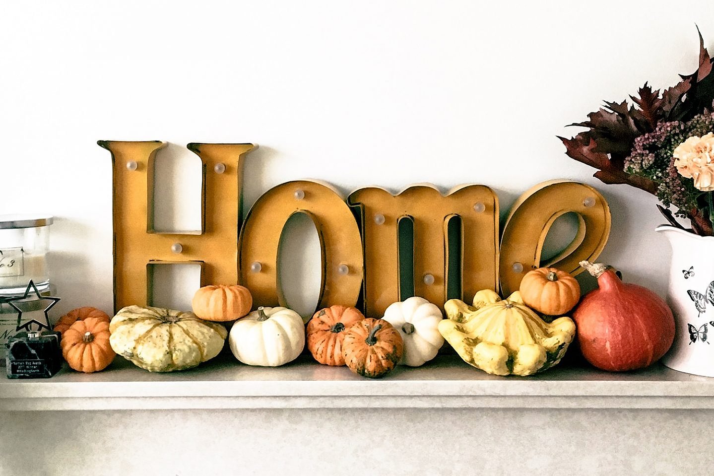 Home sign sits on fireplace surrounded by gourds and pumpkins