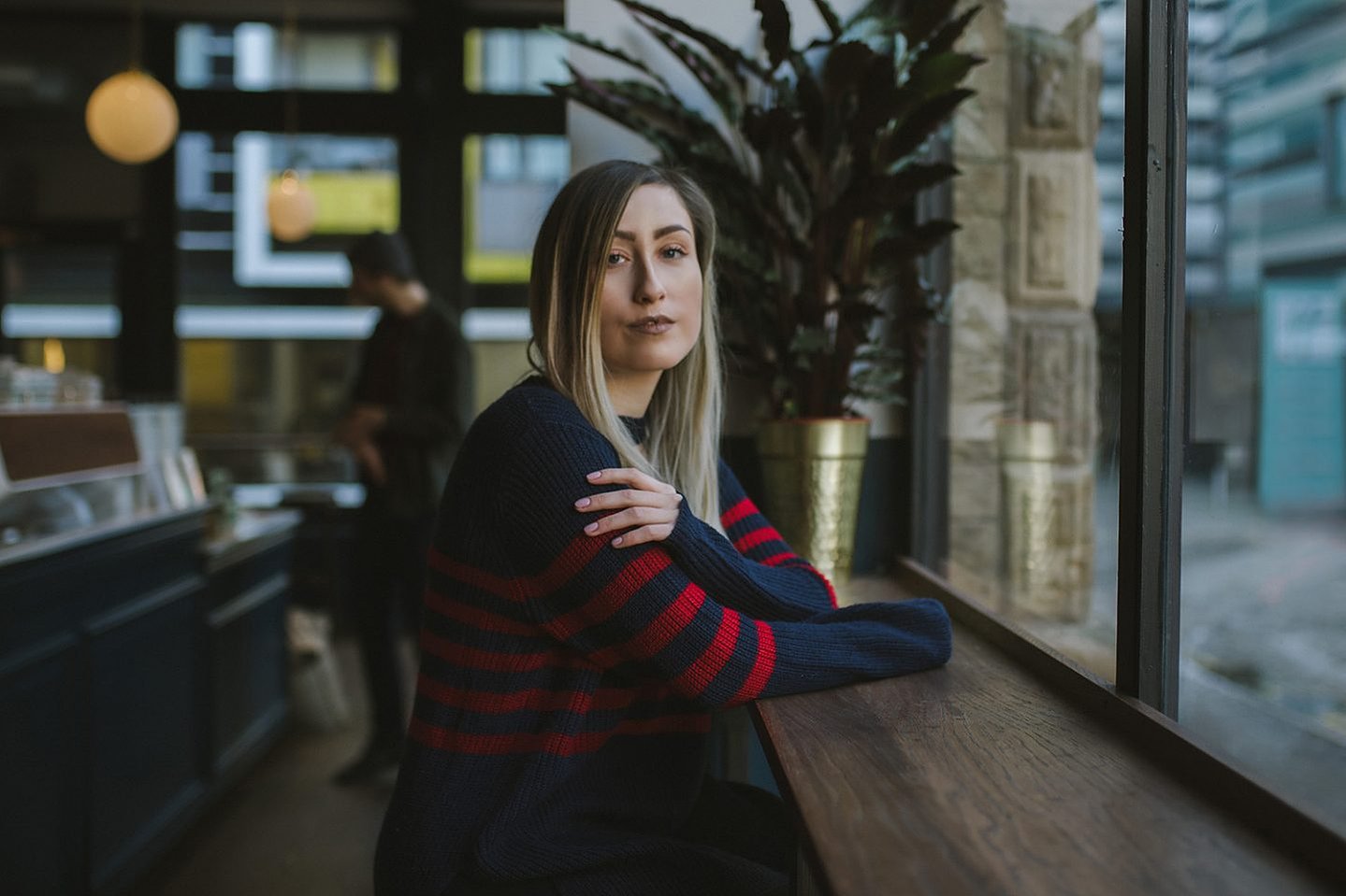 girl sits in coffee bar wearing striped jumper dress, with brown to blonde balayage hair smiling at the camera