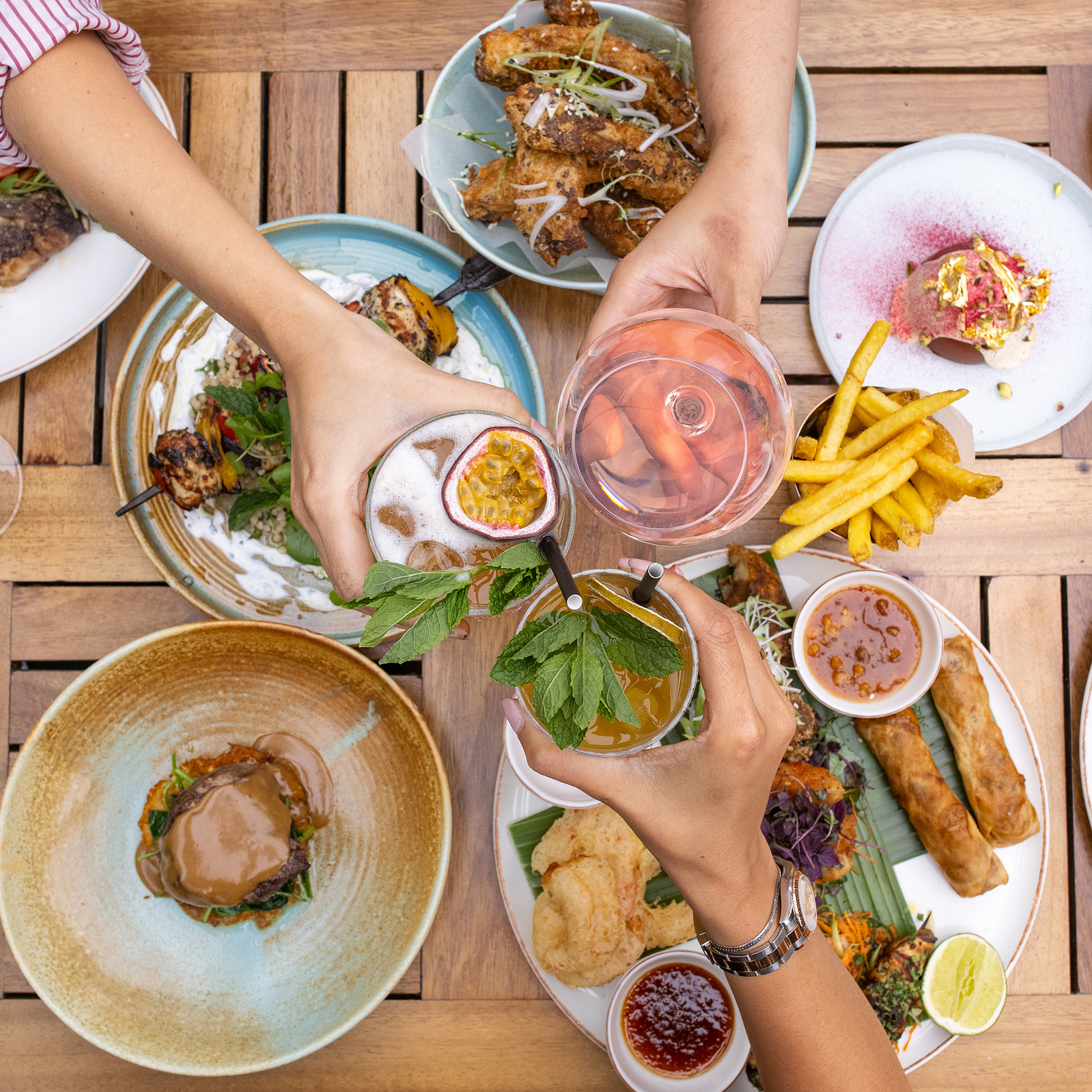 Gather around the table and savor the moment with this lively image from Georgie Glass Photography. This shot captures the essence of social dining, with hands reaching in to enjoy an eclectic mix of sumptuous dishes and refreshing drinks.
