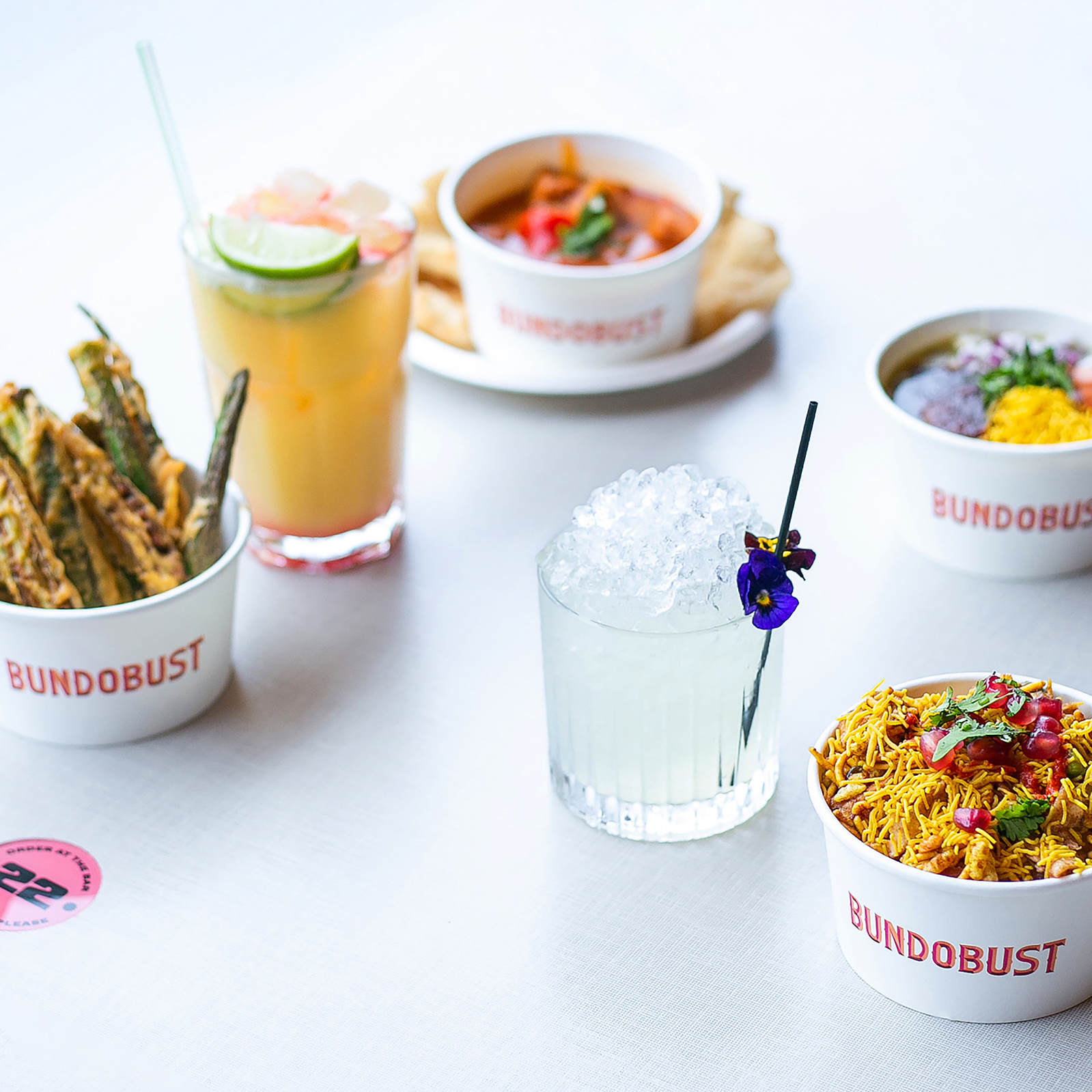Spread of Indian street food and cocktails by Bundobust, with crisp okra fries, vibrant noodle dish, and savory curry, accompanied by a chilled cocktail garnished with a pansy, all captured with the clarity and artistry of Georgie Glass Photography.