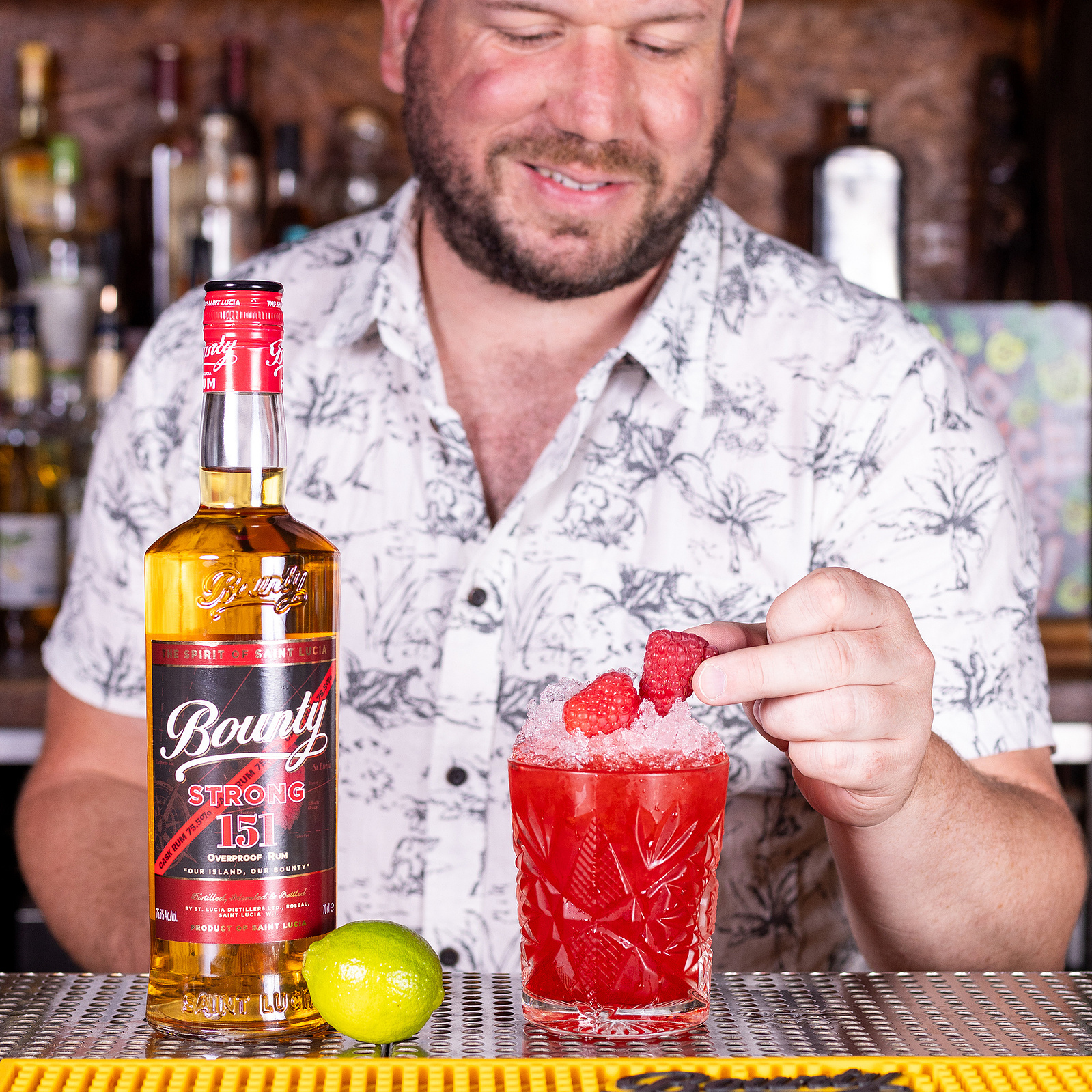 A cheerful bartender garnishing a vibrant raspberry cocktail with fresh fruit, with a bottle of Bounty Strong 151 rum prominently displayed, captured by Georgie Glass Photography, showcasing the lively spirit and rich flavours offered by Bounty Rum.