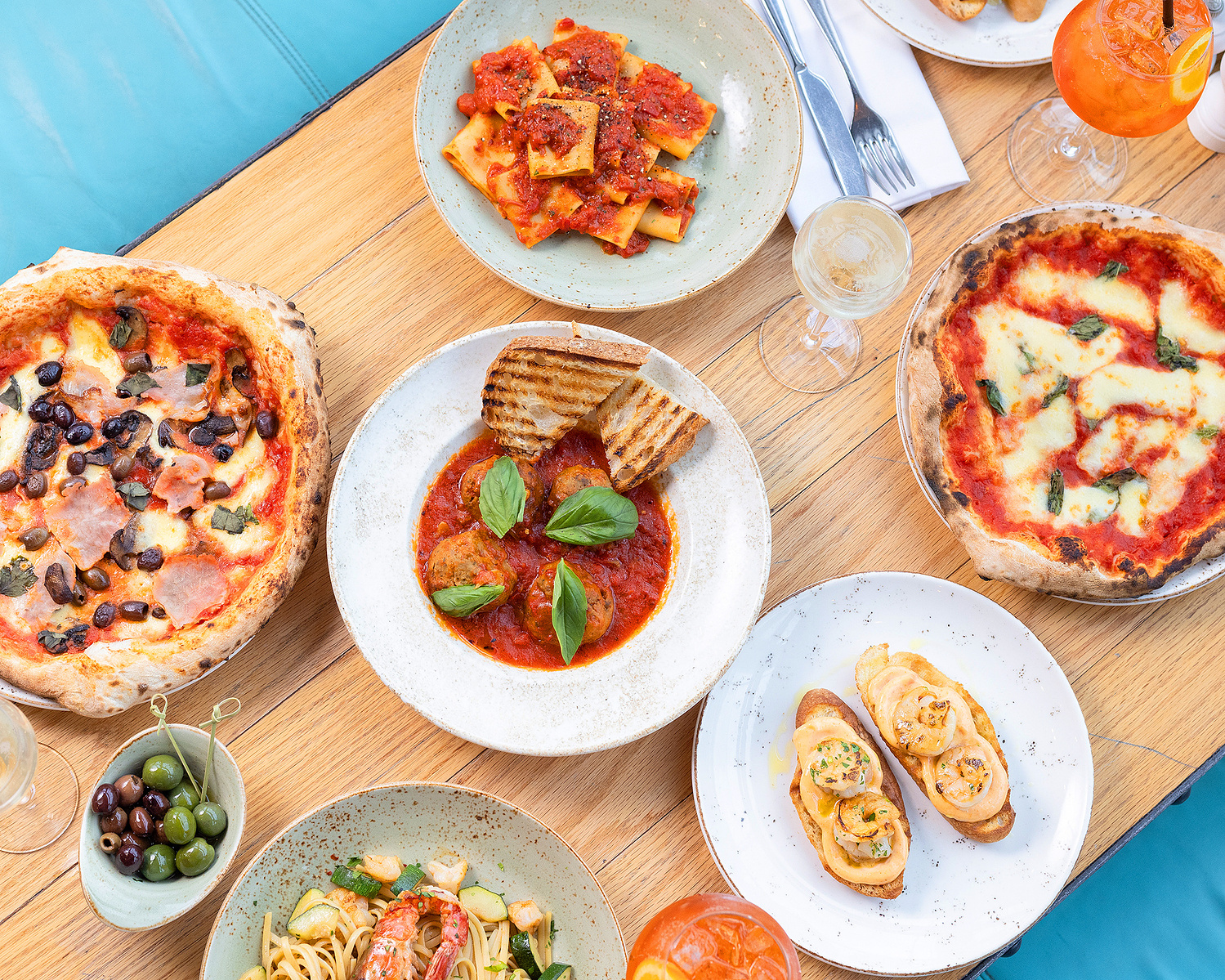 A vibrant array of Italian dishes displayed on a wooden table captured by Georgie Glass Photography, featuring a rustic pizza with olives and ham, a plate of paccheri pasta in tomato sauce, golden toasted panini, and linguine with prawns.