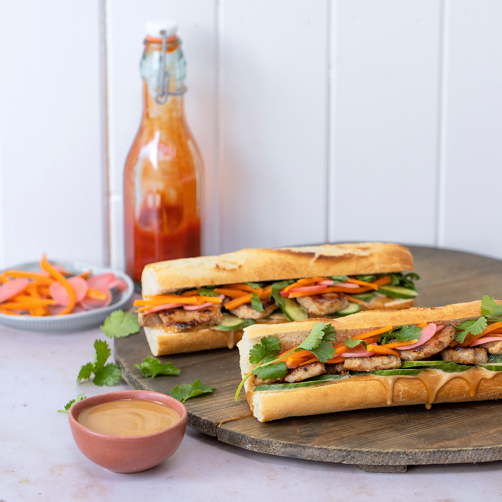 Artistically presented Vietnamese Banh Mi sandwiches on a wooden board, with a side of pickled vegetables, fresh coriander, and a spicy sauce, captured by Georgie Glass Photography.