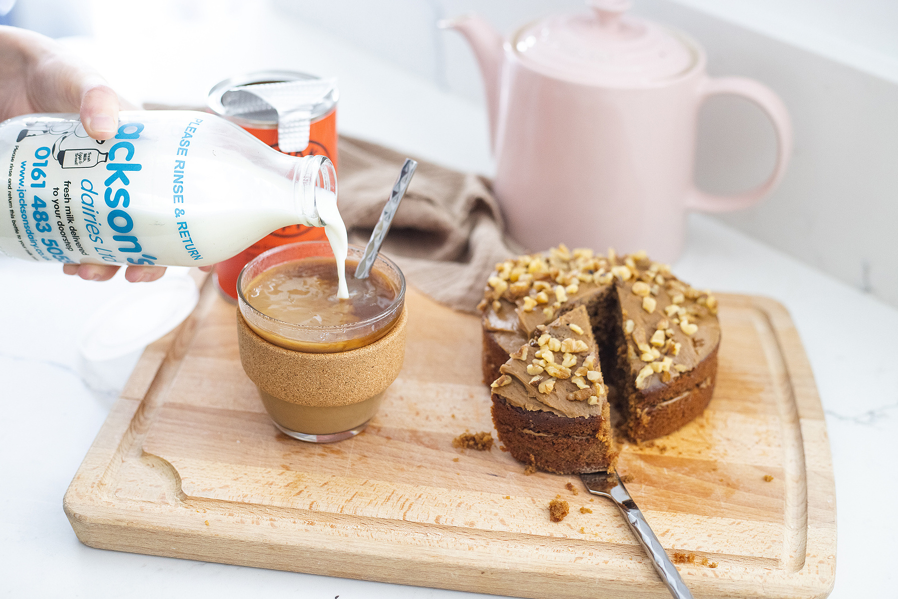 a glass bottle of milk being poured into a coffee on a wooden chopping board next to a cake