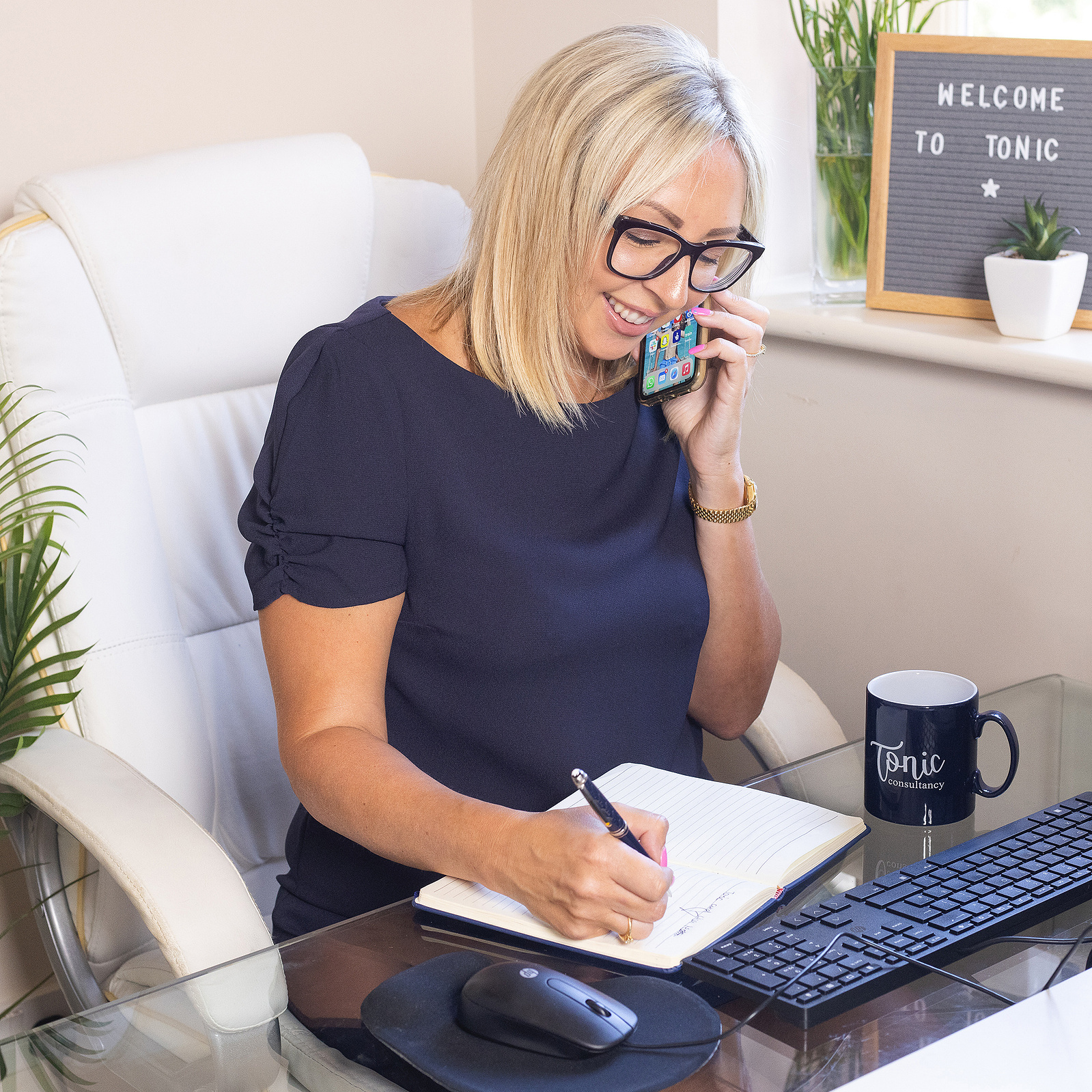 Woman sat at her desk on her mobile phone, writing notes on a note pad. Portrait photography by Georgie Glass, Manchester photographer.