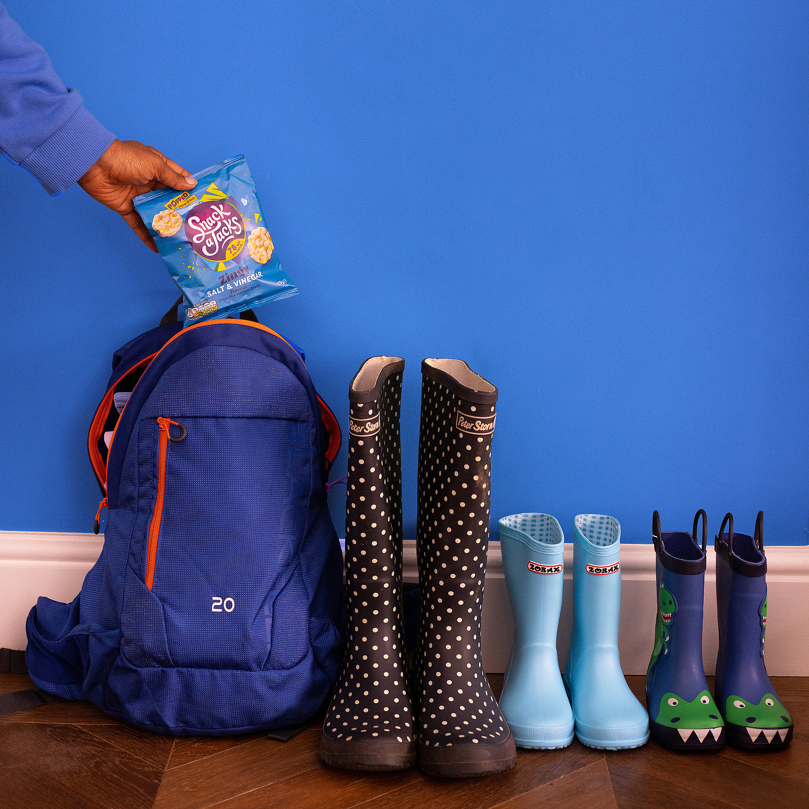 school rucksack on the floor next to 3 pairs of wellington boots. A hand is placing a bag of Snack A Jacks crisps in the rucksack. Product photography by Georgie Glass, Manchester photographer.