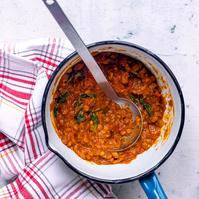 Close-up of a traditional lentil stew with a spoon resting inside the pot, displaying the rich mix of lentils, tomatoes, and greens, presented on a white and red striped kitchen towel, captured by Georgie Glass, Manchester.