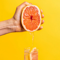 A vibrant and fresh grapefruit half being squeezed by a hand over a glass of sparkling water, set against a bright yellow background, captured by Georgie Glass, food photographer based in Manchester, UK, highlighting the lively burst of citrus.