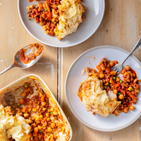 Top-down shot of a rustic shepherd's pie with one serving taken out, revealing a colorful mixture of minced meat, chickpeas, and vegetables beneath mashed potato crust, on a wooden table, by Georgie Glass food photographer from Manchester.