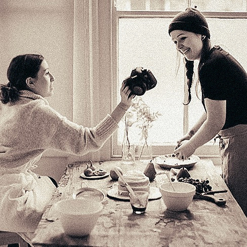 two women laugh and look over images on the back of a camera whilst preparing baked goods as part of a cookery class