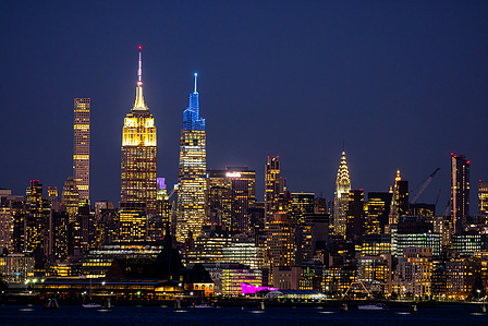 A panoramic view of Lower Manhattan’s skyline at dusk, featuring One World Trade Center and the Brooklyn Bridge reflecting in the calm waters of the East River.