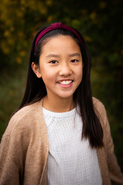 A 12 year old girl poses for a portrait in Bruton Somerset. She's wearing a red hairband, white t shirt and beige cardigan.