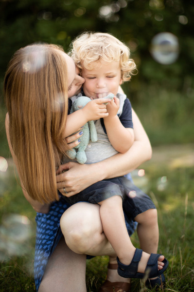 A mother holds her two year old son whilst he sucks his thumb and holds a soft toy. Bubbles are in the foreground. Somerset countryside is blurred in the background.