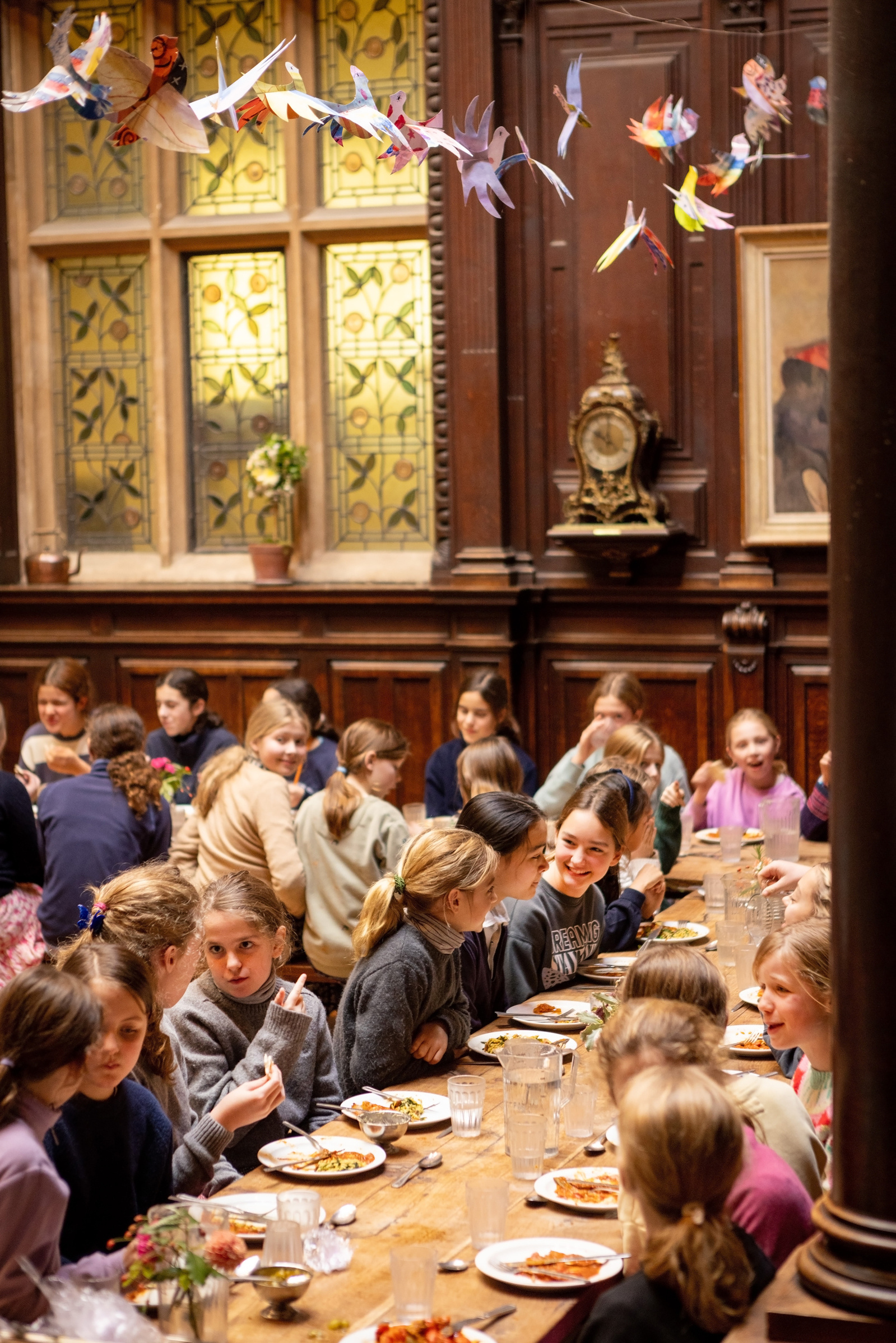 Pupils sit at long tables in the dining room of a grand private school. They are eating and laughing and paper birds hang from the ceiling in front of a grandfather clock.
