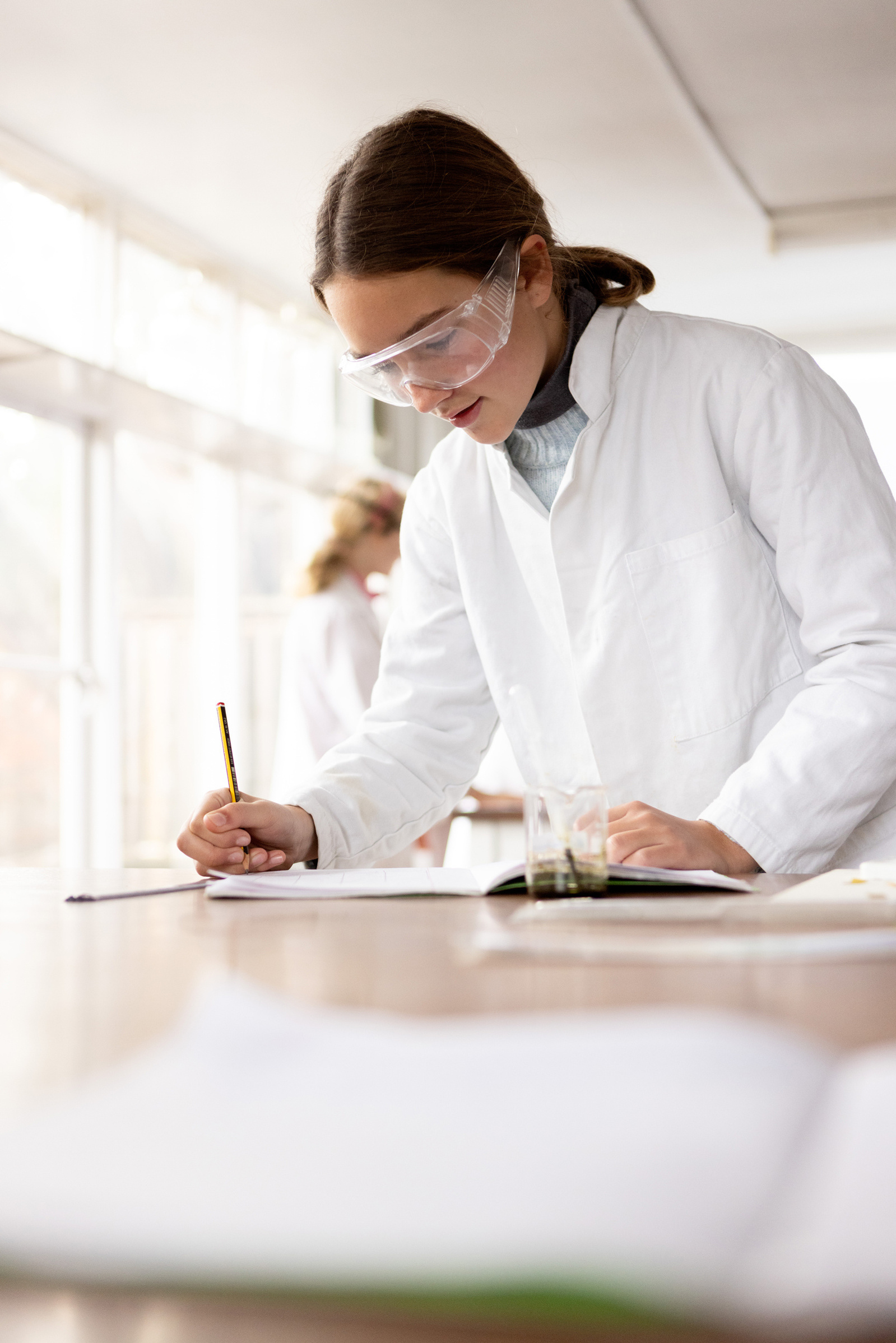 A girl in a lab classroom writes down her findings at the end of the lesson at a private school. She's wearing a white lab coat and science glasses.