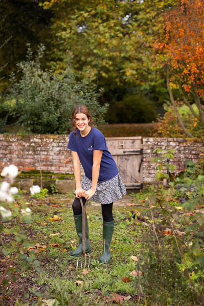 A student stands in a school garden with a garden fork. She is surrounded with autumnal orange and green trees and flowers. She has a wall behind her.