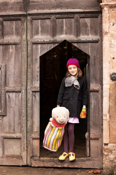 A boarder arrives back at school and stands in the doorway of the school hall with a stripy bag. The bag has her teddy in it.