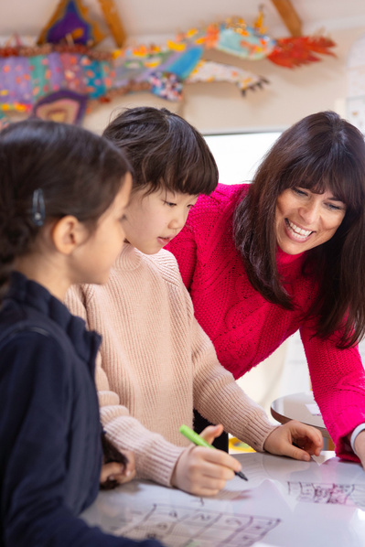 A teacher looks over the shoulder of two young girls who are doing maths sums. They are all smiling and there is a colourful dragon on the classroom wall.
