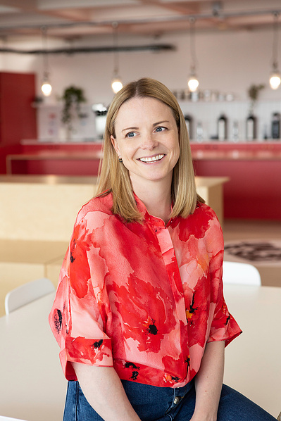 An architect in a red blouse looks off to the left. She is sitting in front of a kitchen in an office in London.