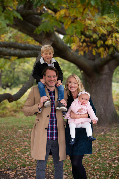 A family of four stand in front of an old tree in Blandford Forest, Dorset. The son is on his Father's shoulders and the Mother is carrying the baby girl. They're all smiling and posing for the camera.