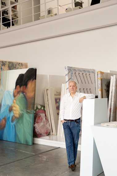 A business man stands in his art studio surrounded by large paintings. He is posing for an environmental portrait wearing a white shirt and jeans and looking straight at the camera. 