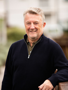 A busines man poses for a portrait outside on a boardwalk in Dorchester. He is wearing a black jumper and shirt.