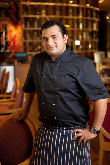 A chef in black with an apron poses for an environmental portrait in front of his restaurant. There are tables and a rack of wine behind him.