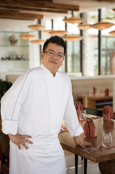 A chef in chef whites stands for an environmental portrait in front of his restaurant. The tables are all laid for service. He is smiling slightly at the camera.