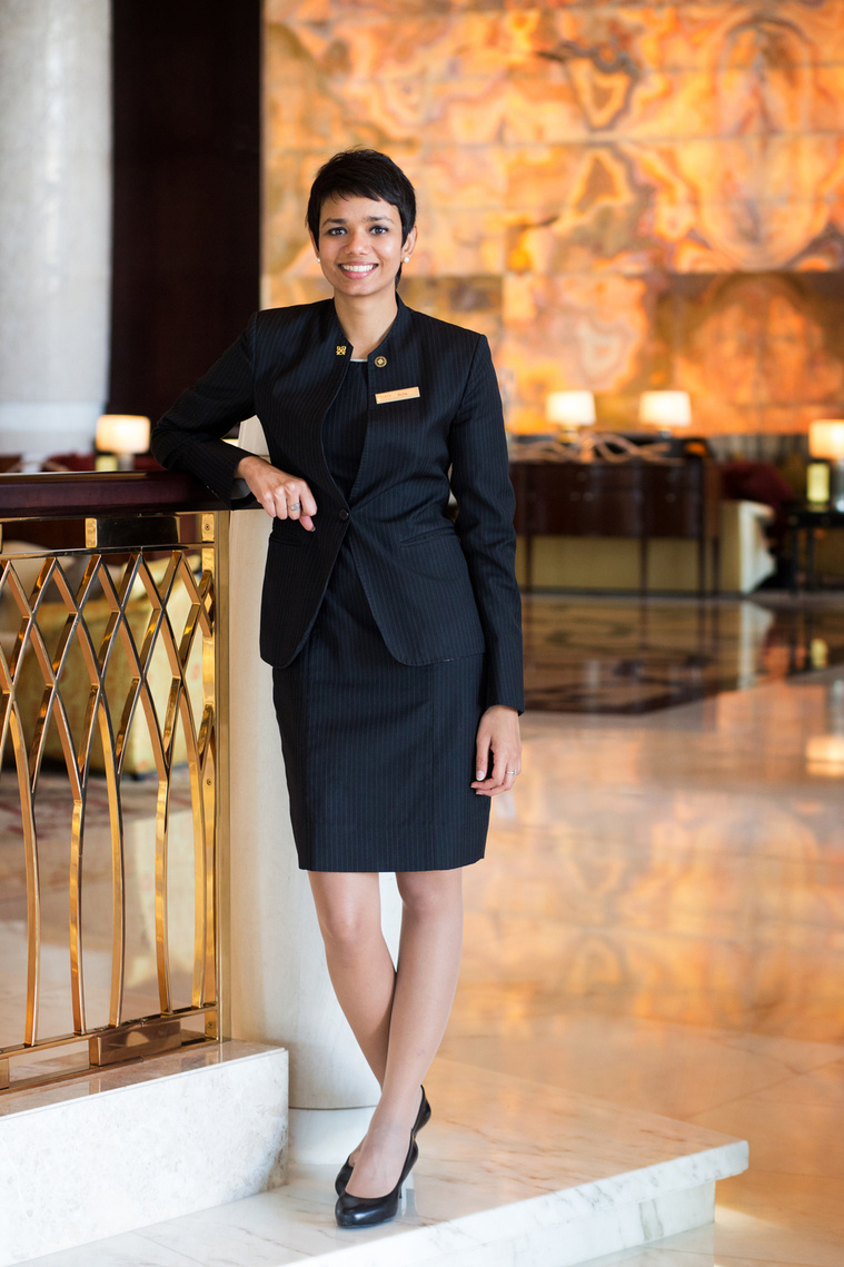 A hotel executive poses for an environmental portrait inside a large hotel reception in London. She is wearing a smart black suit and leaning against a gold balcony.