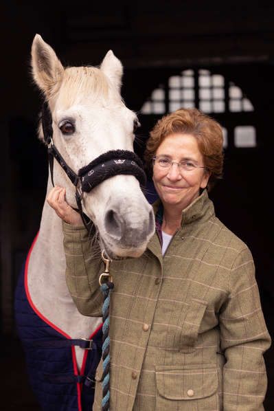 The Headmistress of Hanford School poses for a portrait with her white horse. She is wearing a riding jacket and smiling.