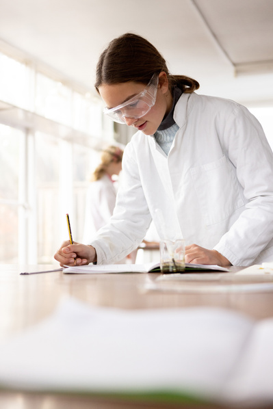 A teenage student in a lab coat in an independent secondary school laboratory in Southampton writes notes. She is in white and the lab is bright and airy. 