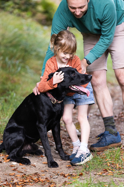 A daughter hugs her black dog, with her Father looking over her shoulder at Blandford Forest, Dorset.