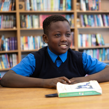 A school boy in uniform smiles at a friend. He's in a school library in front of book shelves sitting at a table with a book in front of him. Photographed at Sunningdale School, Ascot