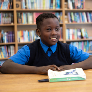 A school boy in uniform smiles at a friend. He's in a school library in front of book shelves sitting at a table with a book in front of him. Photographed at Sunningdale School, Ascot