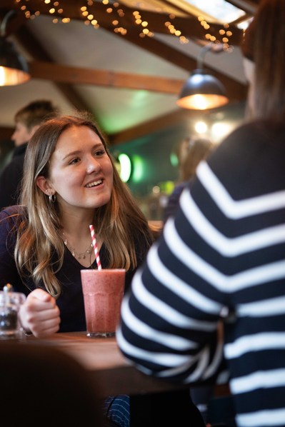 Two female sixth form students at Clayesmore Dorset sit at a table in a cafe with a smoothie chatting.
