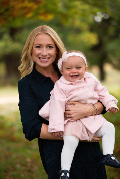 A mother holds her baby girl. They are both smiling at the camera. There's countryside behind them.