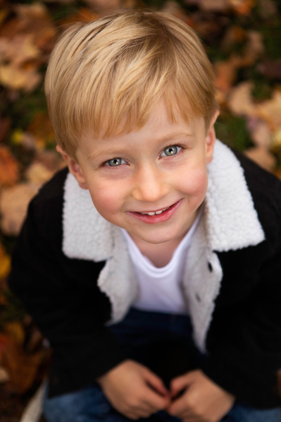 A 5 year old blonde boy looks up at the camera. The shot is taken from above and he is sitting on a pile of leaves in Sherborne.