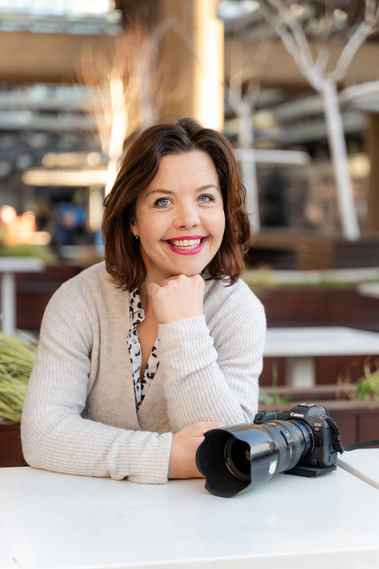 A professional photographer poses for a photograph with her camera. She is outside in Dorchester at a cafe and the background behind her is blurred buildings.