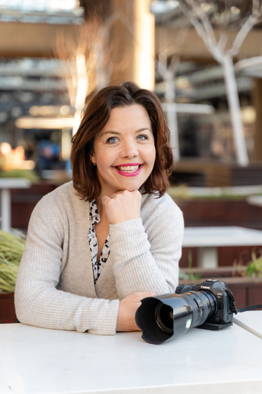A professional photographer poses for a photograph with her camera. She is outside in Dorchester at a cafe and the background behind her is blurred buildings.
