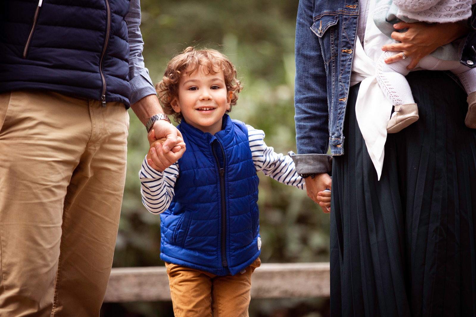 A young boy holding both his parents hands smiles and looks straight at the camera on a boardwalk in Upton Park, Poole, Dorset