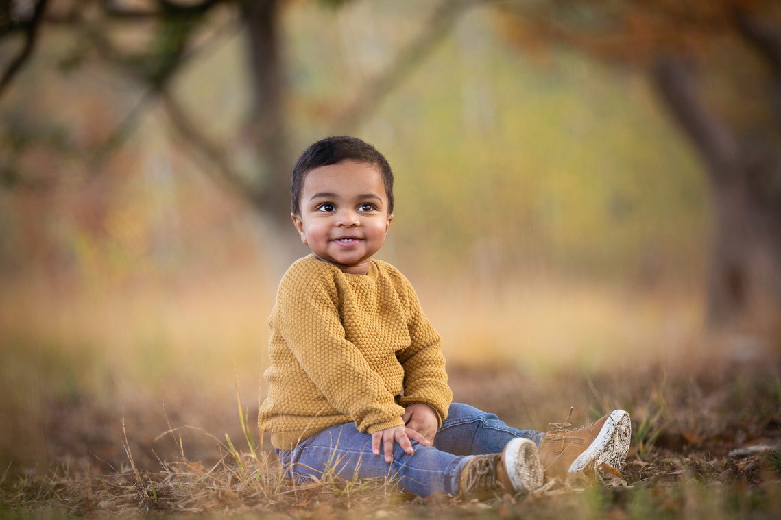 A very cute boy poses for a photo and is looking just to the right. He is sitting down on some dry yellow grass and there are woods behind him. He is grinning and the background is very blurred.
