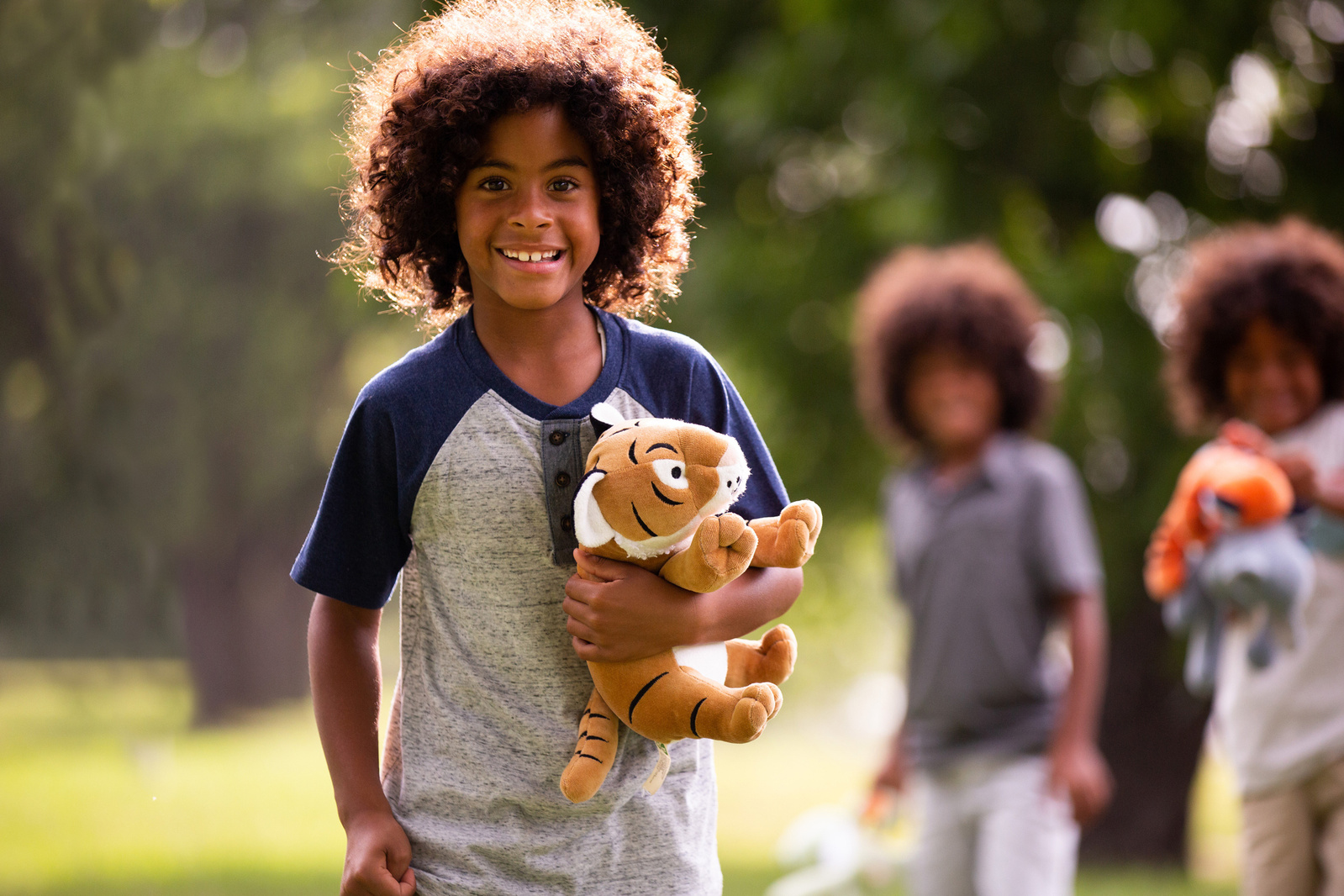 A boy smiles at the camera whilst holding a tiger soft toy. His two brothers are blurred in the background. He's in Upton Country Park, Poole.