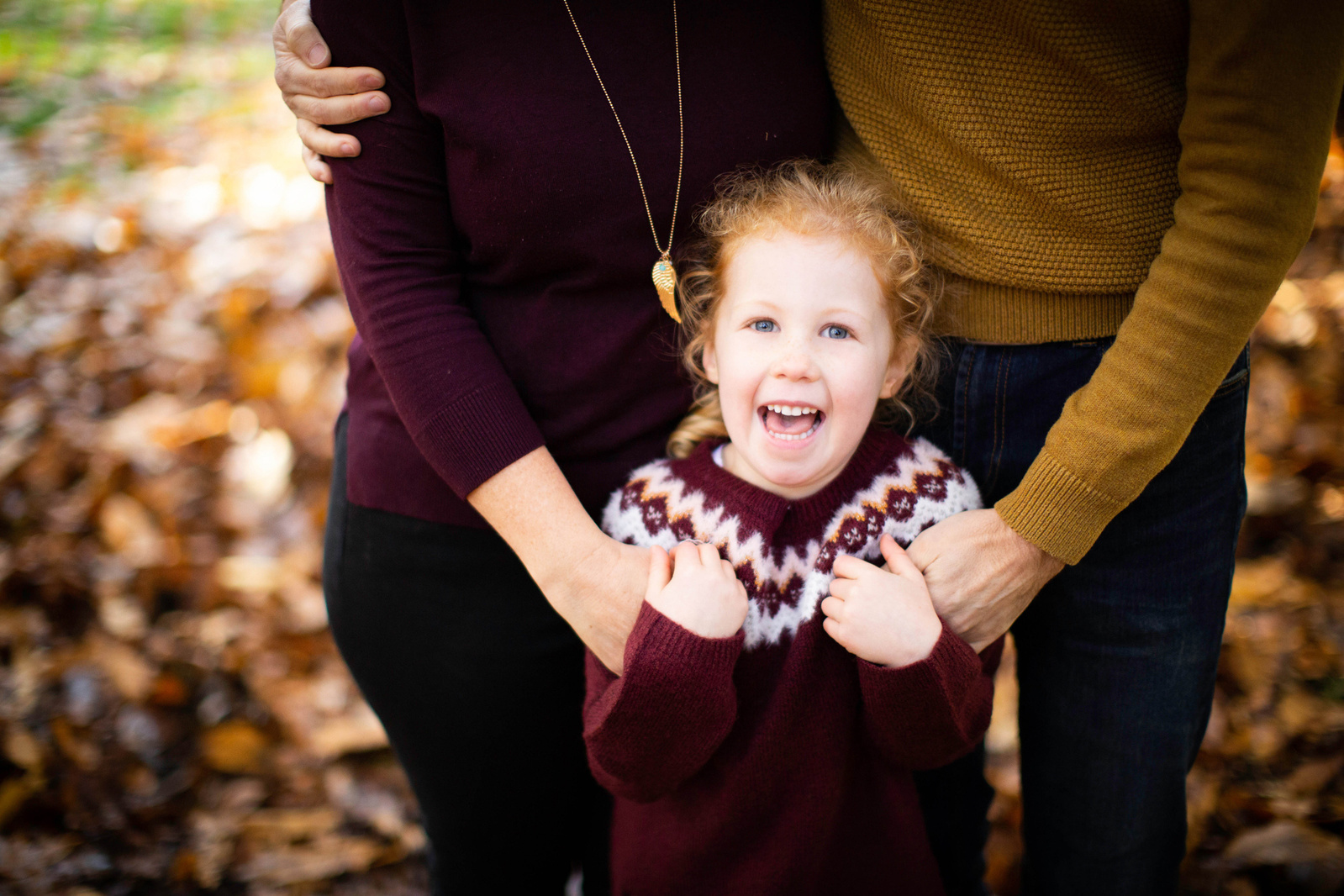 A girl stands in front of her parents. We can't see her parents heads. She is laughing. There are autumnal leaves on the ground in the background. They're in Blandford Forest.