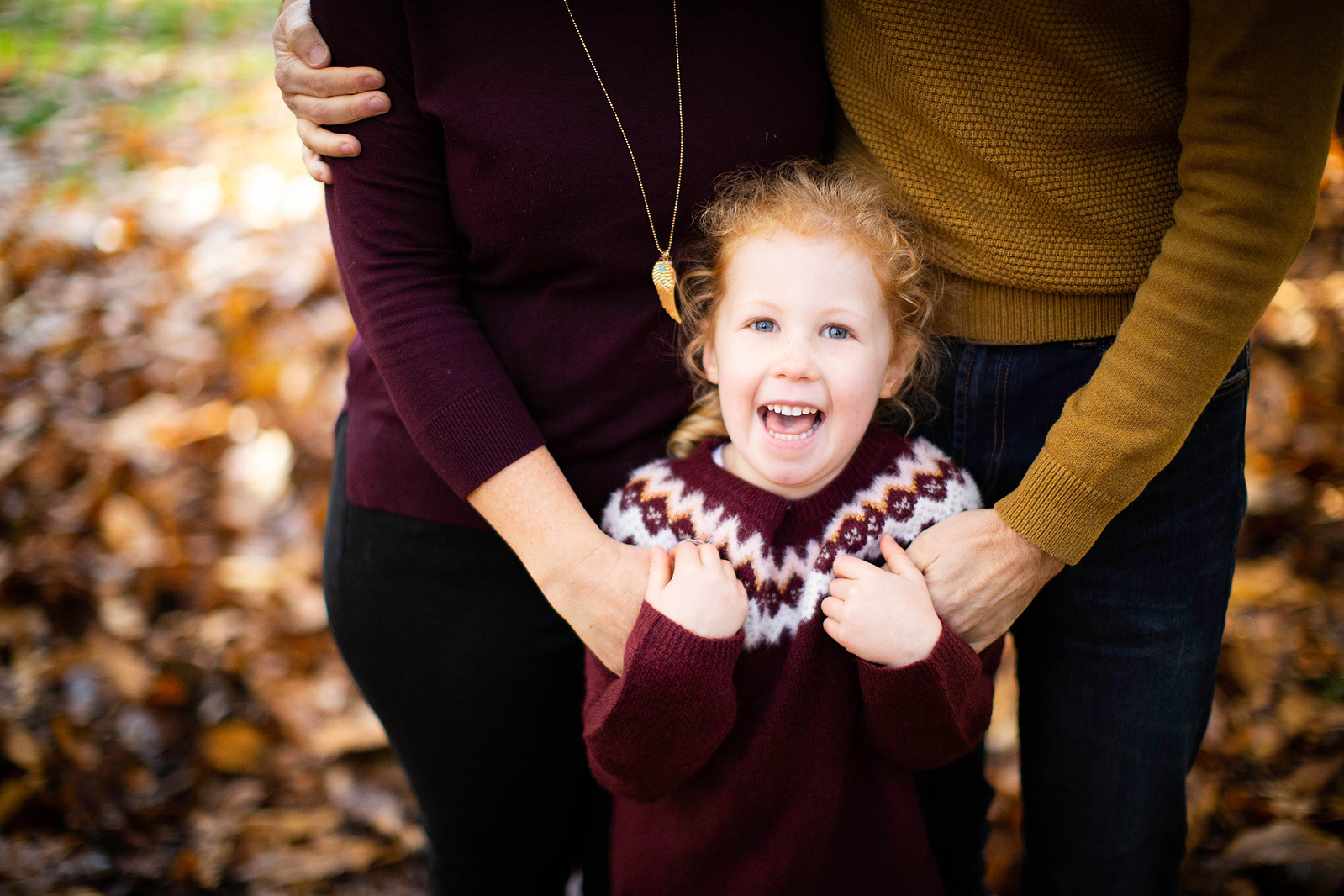 A girl stands in front of her parents. We can't see her parents heads. She is laughing. There are autumnal leaves on the ground in the background. They're in Blandford Forest.